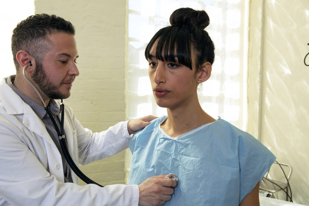 a_transgender_woman_in_a_hospital_gown_being_treated_by_a_doctor_a_transgender_man