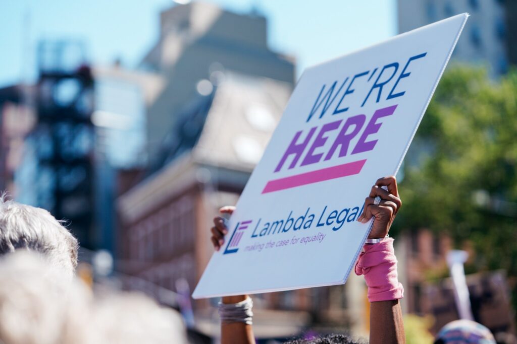 Person holds up white sign that reads "We're Here" with Lambda Legal logo